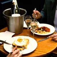 A bottle of champagne in an ice bucket, two full glasses and two main meals sit on a table at La Buvette