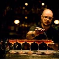 A bartender drops the finishing touches into 5 cocktails lined in a row at Maybe Mae 