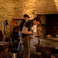 A forgeron holds a hammer ready to hit a metallic item and an older man stands in front of braiser.