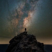 Someone stands on the top of a large rock, surrounded by stars and the Milky Way overhead 