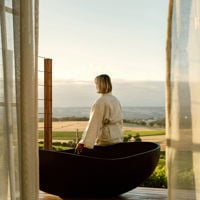 Draping curtains frame someone who sits on the edge of the black bath in their robe looking towards the vines