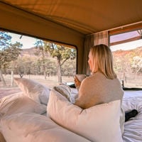 a woman sits propped up against some pillows with a cup of coffee in ahnd as she looks out of her canvas tent