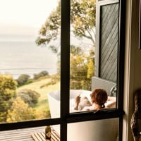Through the window on the you see someone in the bath which looks out over the green foliage to the sea 