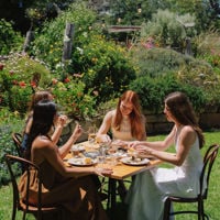 a group of friends sit around an outdoor table, within a garden, enjoying lunch