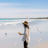woman in a sun hat laughs at the beach with a scroll in hand, seagulls flying around her in the air